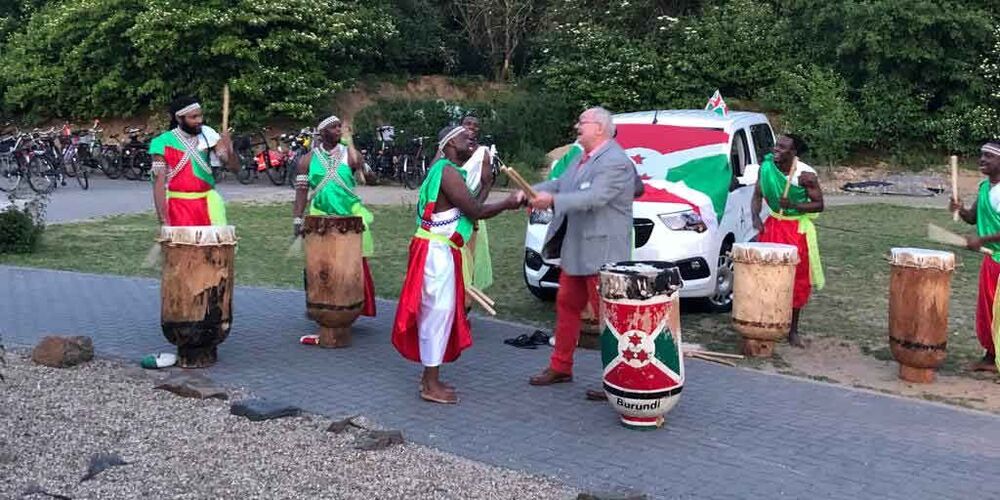 Ein Auto vor dem einige Männer mit Trommeln und afrikanischer Kleidung stehen. Ein weiterer Mann im Anzug steht auch vor diesem und wird von einem der anderen Männern begrüßt.
